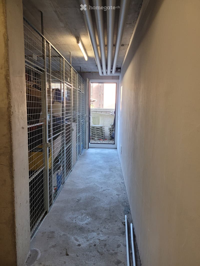Concrete hallway with metal cage on left, open doorway leading outside, stacks of materials visible beyond doorway.