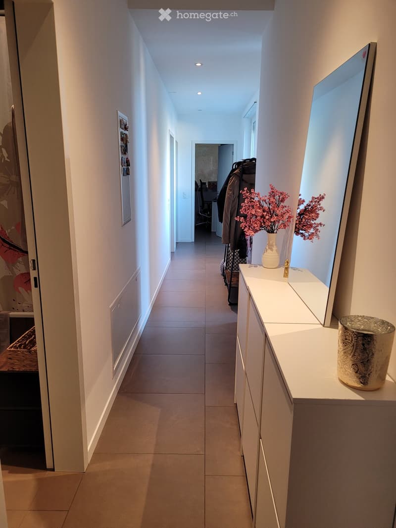 Modern hallway with tiled flooring, white walls, recessed lighting, and a small console table with a vase of pink flowers and a decorative item.