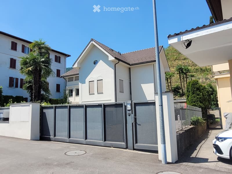 A modern two-story house with white exterior walls, grey tiled roof, and multiple windows. The property features a gated entrance with a security camera and a palm tree in the background.