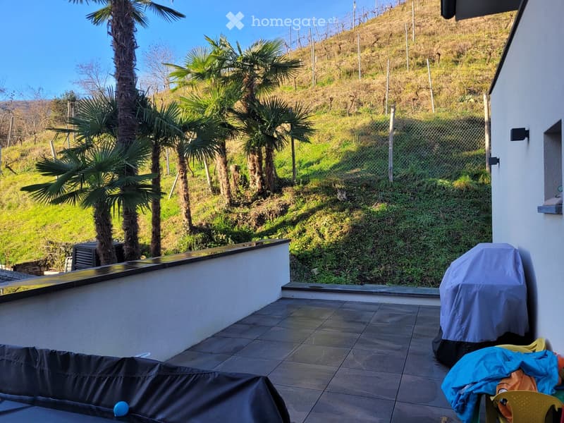 Balcony with tiled floor, covered grill, and outdoor furniture, overlooking a landscaped hillside with palm trees and a fence.