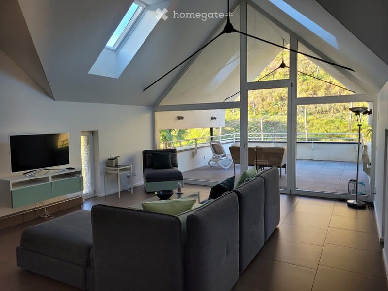Modern living room with large windows and a balcony, featuring a grey sectional sofa, coffee table, TV stand, and additional seating area.
