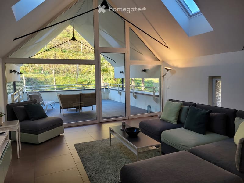 Modern living room with large glass doors opening to a balcony, featuring a sectional sofa, coffee table, and a view of greenery.