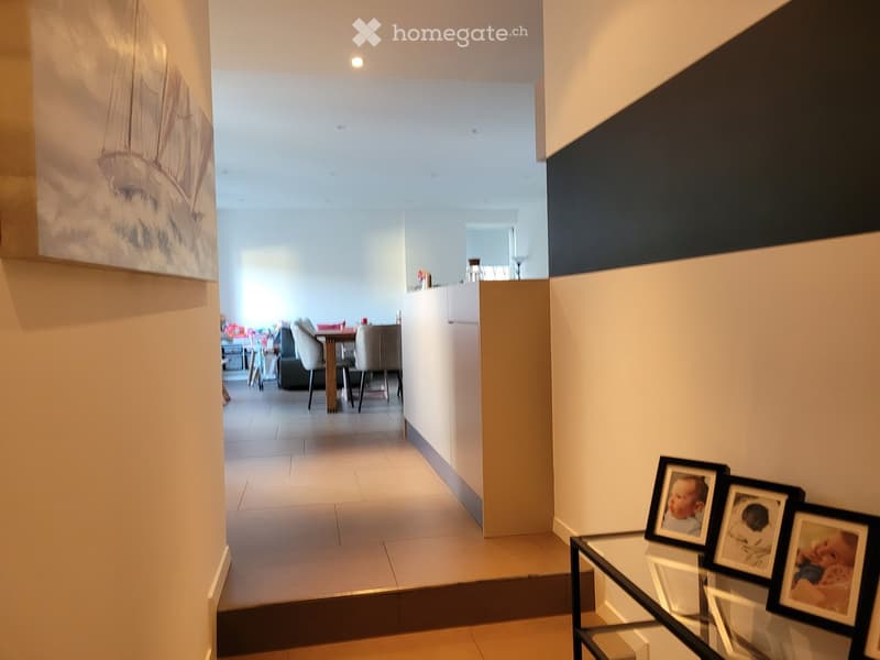 Modern hallway with tiled flooring, white walls, and a glass coffee table with framed photos. The hallway leads to a dining area with a table, chairs, and a couch.