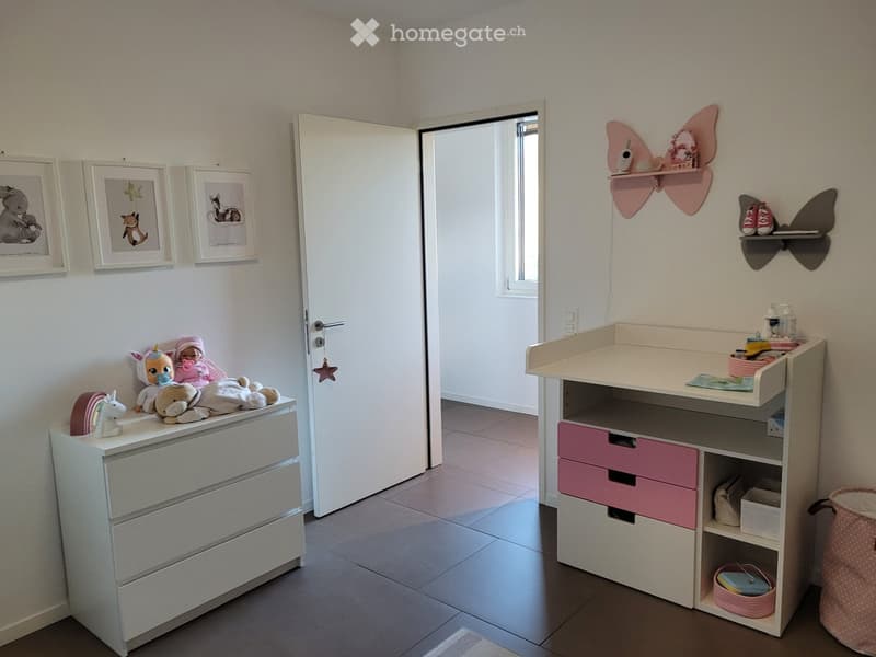 Brightly colored children's bedroom with a changing table, drawer unit, and various toys. The room features framed pictures on the wall and a shelf with additional toys and items.