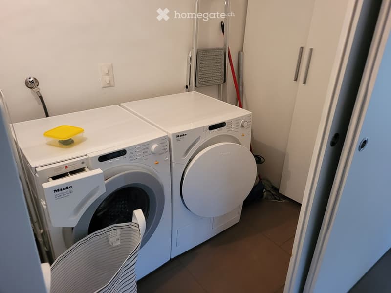 A small bathroom with a washing machine and a dishwasher, white tiled walls, and a striped towel hanging on the side.