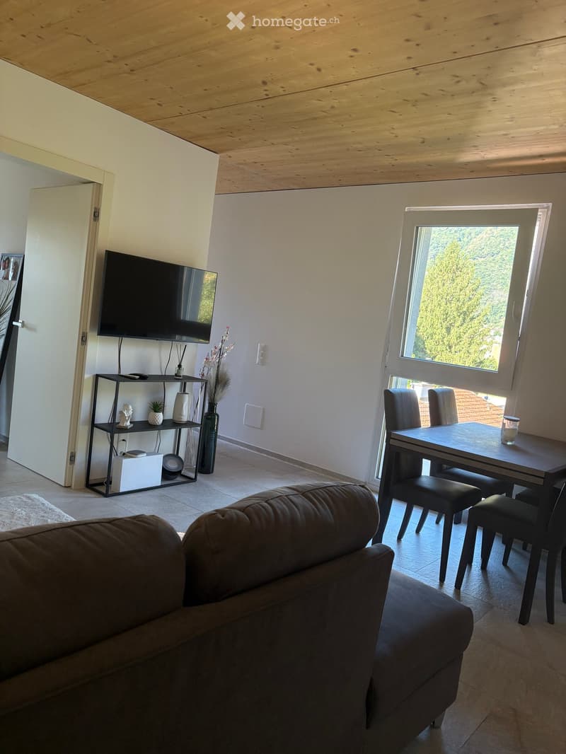 Modern living room with a brown leather sofa, a black dining table with four chairs, a wall-mounted TV, a black shelf with decorative items, and a large window with a view of greenery.