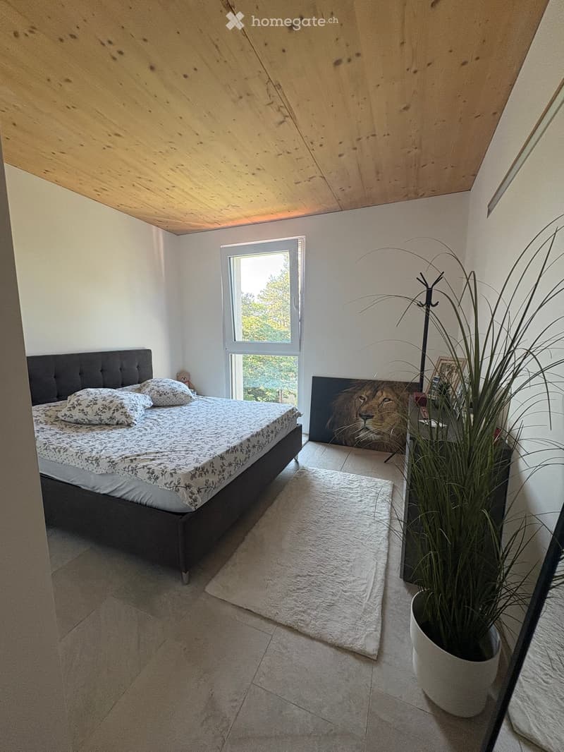 Bright bedroom with a wooden slanted ceiling, large window, bed with patterned bedding, white rug, potted plant, and a picture of a lion on the wall.
