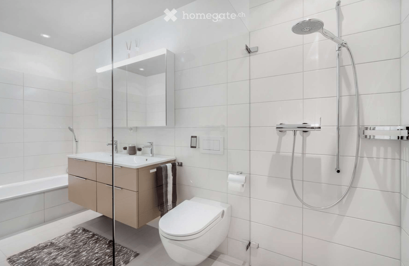 Modern bathroom with white tiles, a glass-enclosed shower, a sink with a cabinet, a toilet, and a bathtub. The space includes a mirror, towel, and light switch.