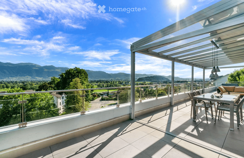 A modern balcony with a glass railing, a covered seating area, and a scenic mountain view.