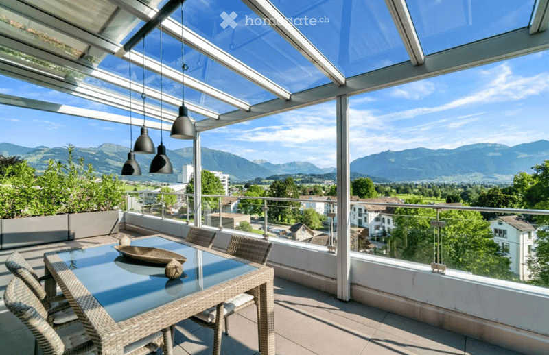 A modern glass-enclosed balcony with a scenic mountain view, featuring a glass table and wicker chairs.