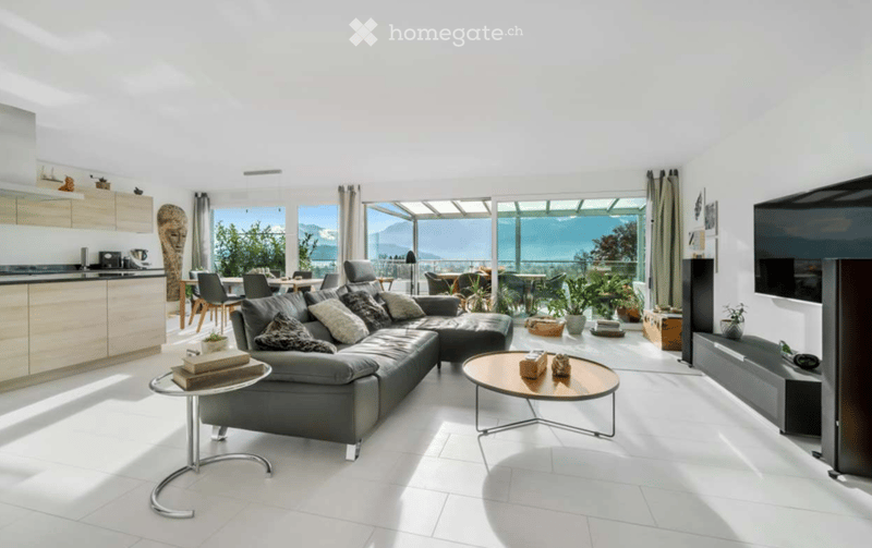Bright, modern living space with white walls, white tiled flooring, and large windows offering a scenic view. Features a grey sectional sofa, coffee tables, a TV stand, kitchen area with cabinets, and a dining area with chairs.
