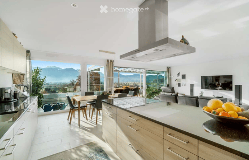 Open-plan kitchen and living area with modern white cabinets, a central island, a dining table with chairs, a sofa, and a television. Large windows offer a scenic mountain view.