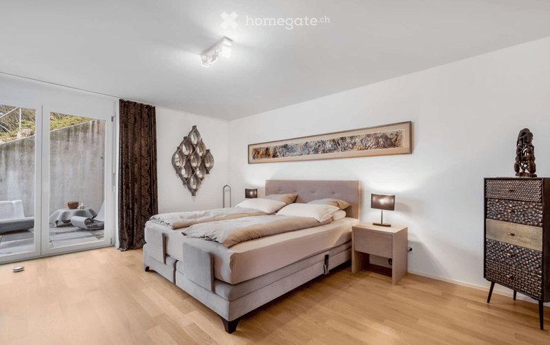 Modern bedroom with a large bed, nightstand with lamp, dresser, wall-mounted mirror, and artwork. Large windows offer a view of the outdoor area.
