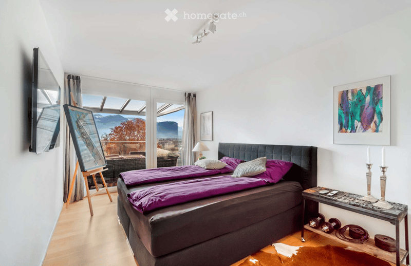 Bright bedroom with a large bed, nightstand, lamp, mirror, picture frame, and a view of the mountains through large windows.