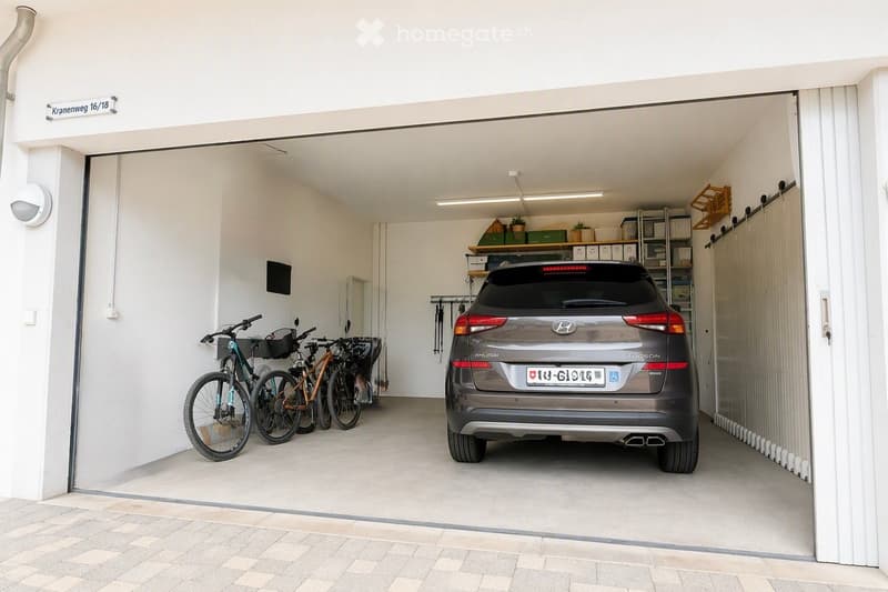 Clean, well-lit garage with a parked car, bicycles, and storage shelves with boxes and potted plants.