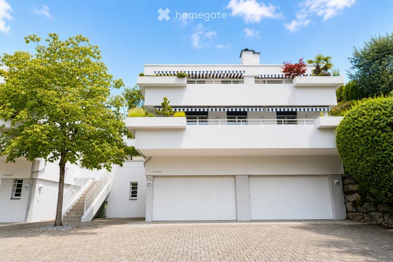 Modern two-story white house with large garage, raised ground floor, and balcony terrace with greenery.