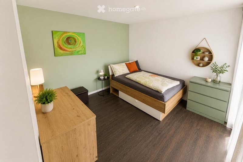 Cozy bedroom with a wooden bed, nightstand, lamp, and decorative plants. Features a green accent wall, a modern painting, and a hanging shelf with small decorative items.