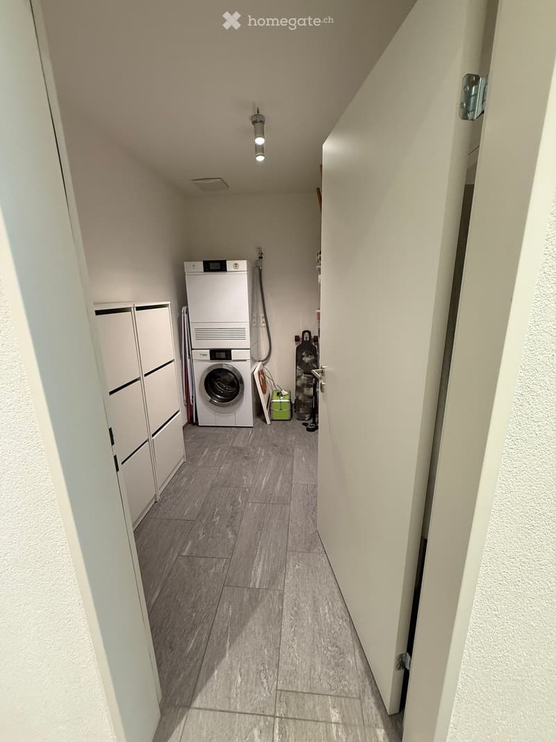 A narrow hallway with a tiled floor, white walls, and a ceiling light. A washing machine is visible in an adjacent room.