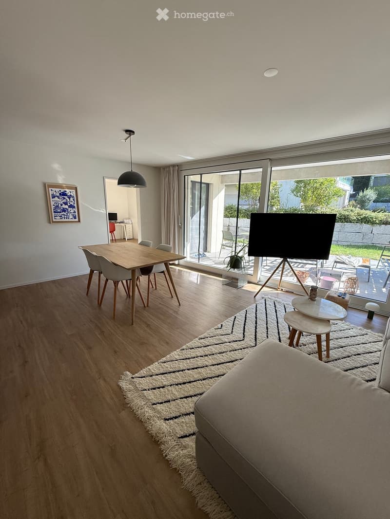 Modern living room with wooden flooring, large windows, a sofa, coffee table, and a dining area with a table and chairs.