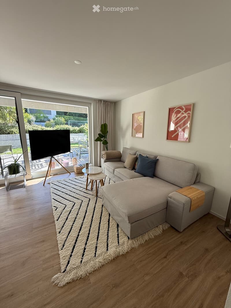 Modern living room with a sectional sofa, coffee table, small stool, television on a stand, and a large area rug. The room features large windows with curtains and a view of a garden.