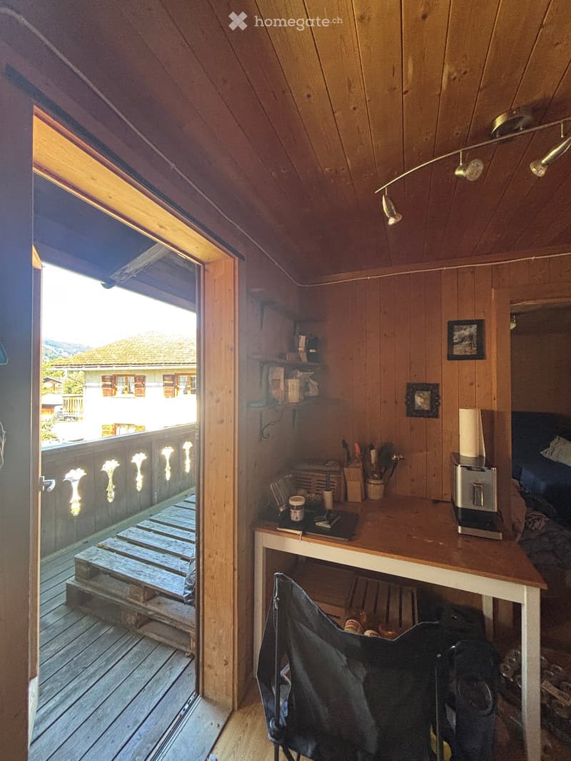 Cozy attic room with wooden walls and ceiling, featuring a small desk, chair, and shelves. An open door leads to a wooden deck with a scenic view.