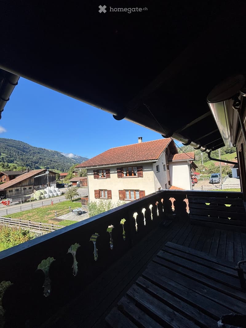 Wooden balcony with railing overlooking a rural village and mountains, with a few parked cars and a small table visible in the distance.