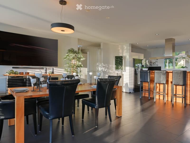 Modern dining room with a wooden dining table, black leather chairs, and grey bar stools. A large flat-screen TV is mounted on the wall, and a hanging pendant light illuminates the space. The room features a sleek, contemporary design with a mix of dark a