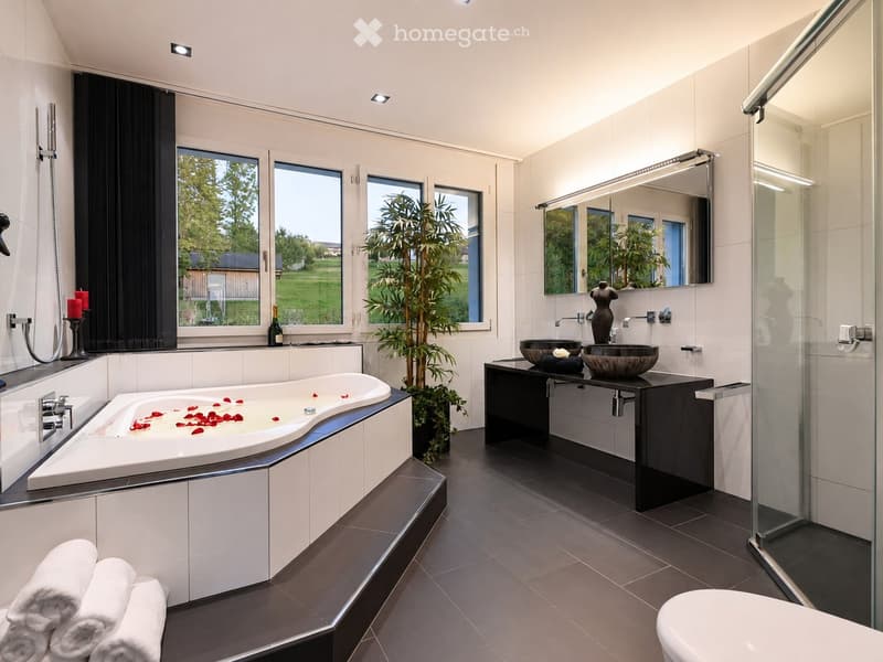 Modern bathroom with a large bathtub, double sink vanity, large mirror, and a glass-enclosed shower. Features include a potted plant, candles, and a view of greenery through large windows.