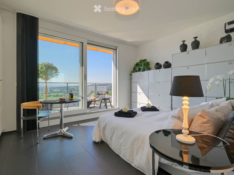 Modern bedroom with a large bed, nightstand, lamp, chair, table, and a view of the landscape through large windows. The room features a sleek, contemporary design with a mix of dark and light tones.