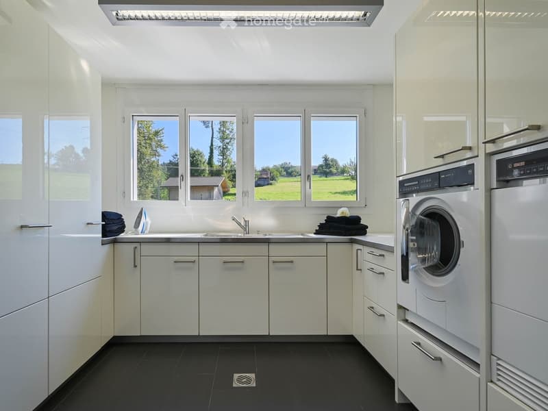 Modern bathroom with white cabinets, black tiled floor, large windows, sink, washing machine, and towels.
