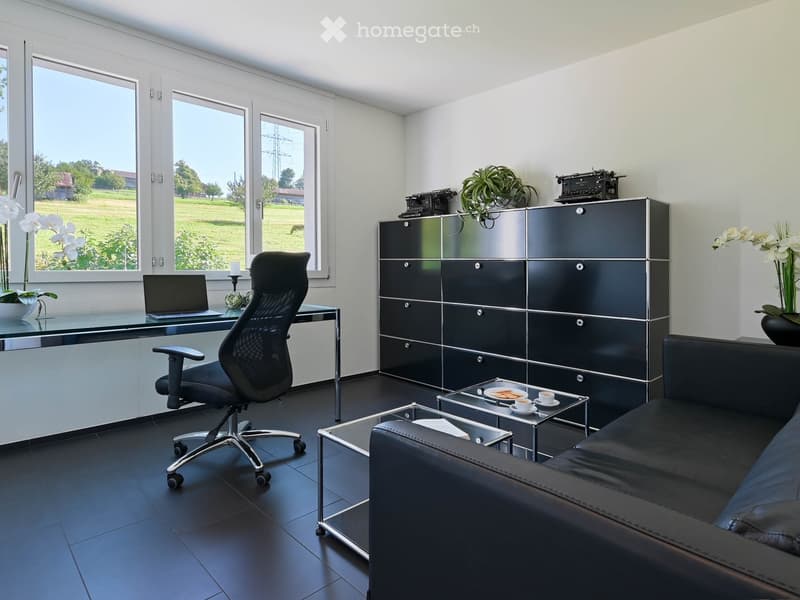 Modern, well-lit room with large windows, black leather sofa, glass coffee table, ergonomic chair, laptop on desk, and decorative items including a typewriter and potted plants.