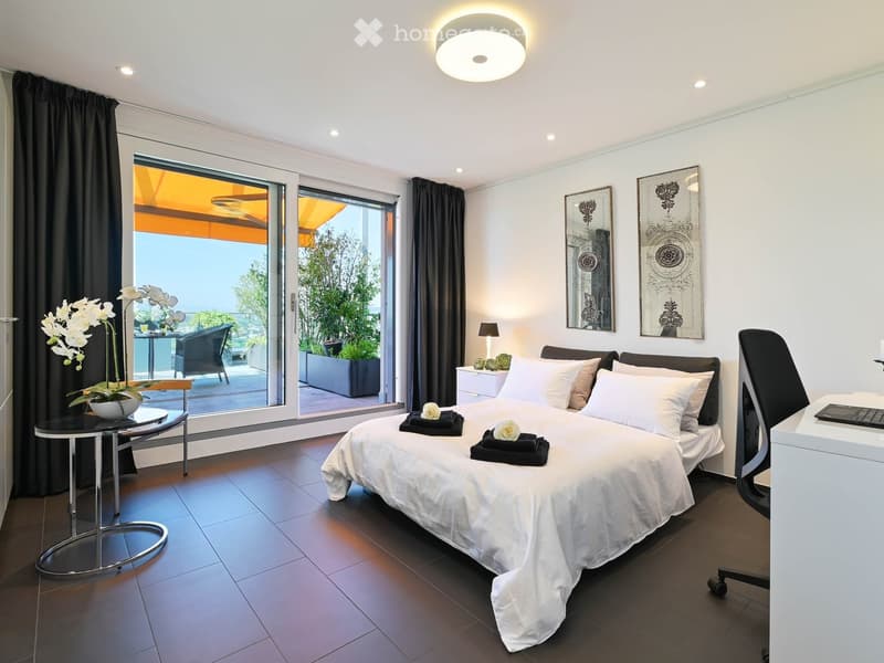 Modern bedroom with a large bed, white linens, decorative pillows, a nightstand with a lamp, a round side table with a flower arrangement, and a large window with a view of a balcony area.
