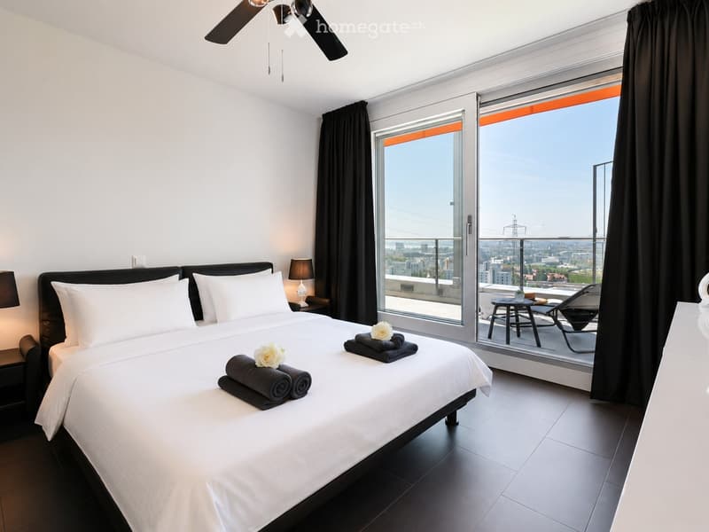 Modern bedroom with a large bed, nightstands, lamps, and a view of the city through large windows.