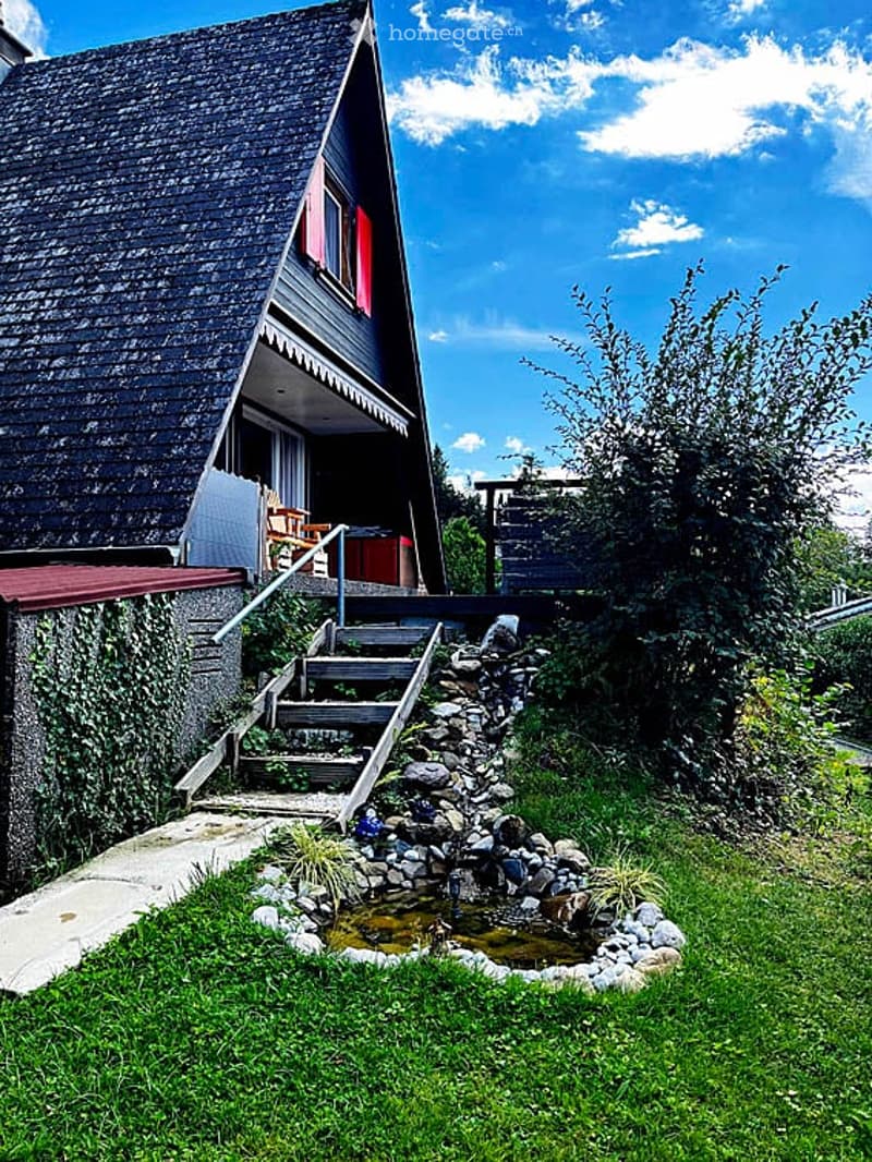 A modern house with a dark wooden roof and red shutters, surrounded by a well-maintained garden featuring a small pond with a stone waterfall, steps leading up to the entrance, and a wooden deck area.