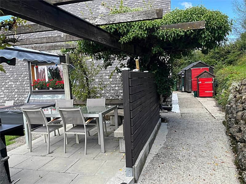 Outdoor patio area with a dining table and chairs, covered by a pergola, surrounded by greenery and a stone pathway leading to a red shed.