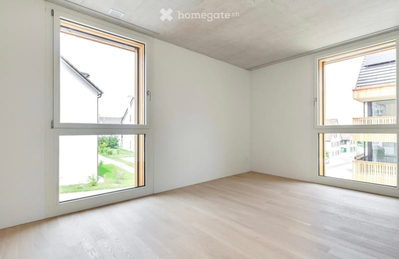 Empty room with light-colored wooden flooring, white walls, and large windows offering views of the surrounding area.