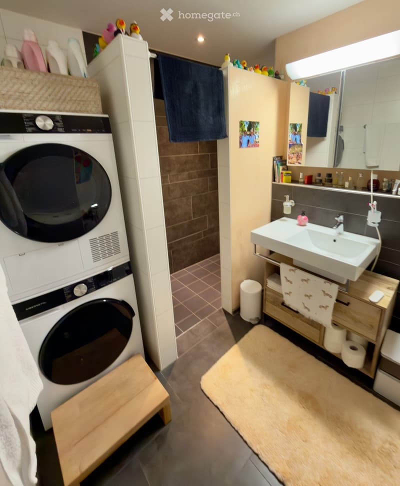 Bathroom with a sink, mirror, cabinet, washing machine, and various personal items. Includes a wooden stool, towel, and bath mat.