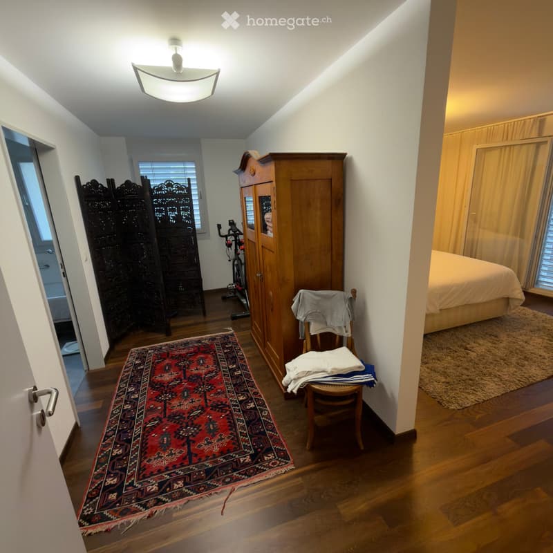 A furnished room with wooden flooring, a large red and black patterned rug, a wooden wardrobe, a bicycle, a screen, a bed, and a chair with folded clothes on it.