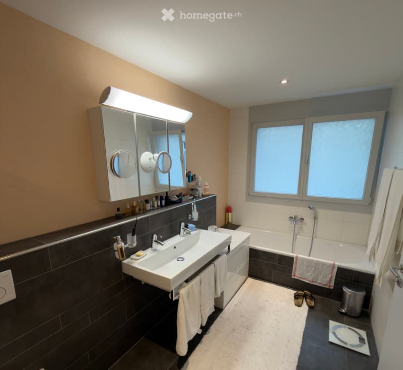 Modern bathroom with a double sink, bathtub, large mirror with built-in lighting, and various toiletries. The space features dark tiles and a white towel on the edge of the bathtub.