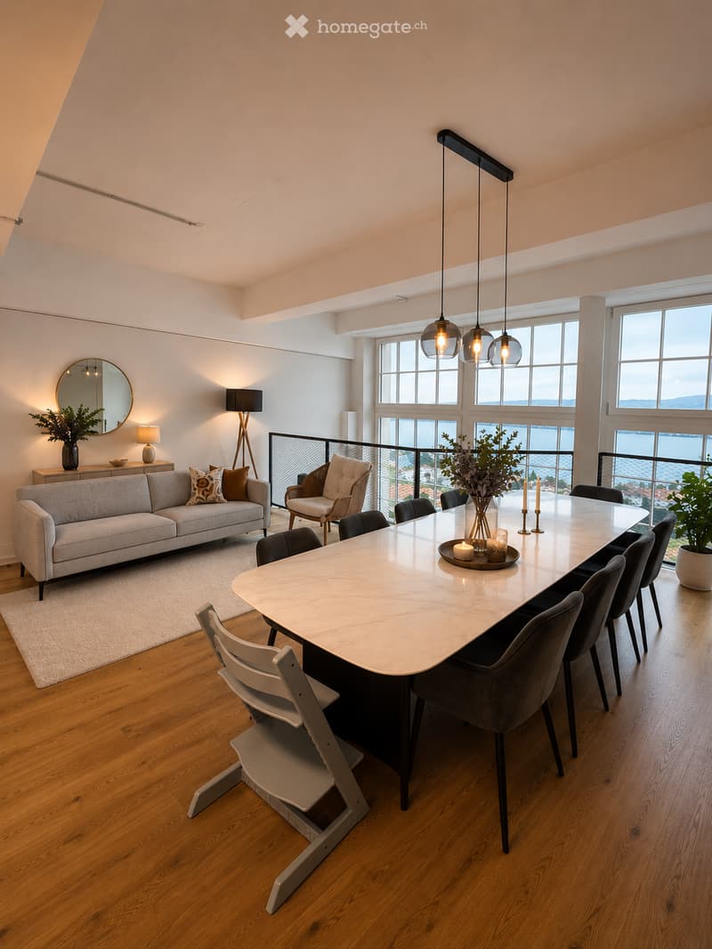 Open-concept living and dining area with a grey sofa, coffee table, dining table with chairs, pendant lights, and a view of the water through large windows and a balcony.