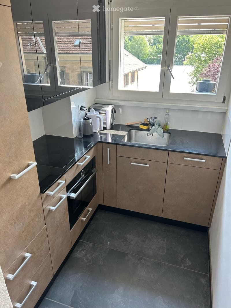 Contemporary kitchen, light wood cabinets, black granite countertops, gas stove, stainless steel sink, window with shutters, modern appliances, and a view of greenery outside
