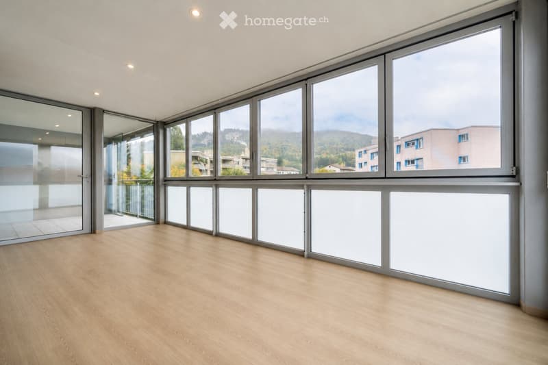 Empty room with large windows and wooden flooring, featuring a modern design and a view of the surrounding area.