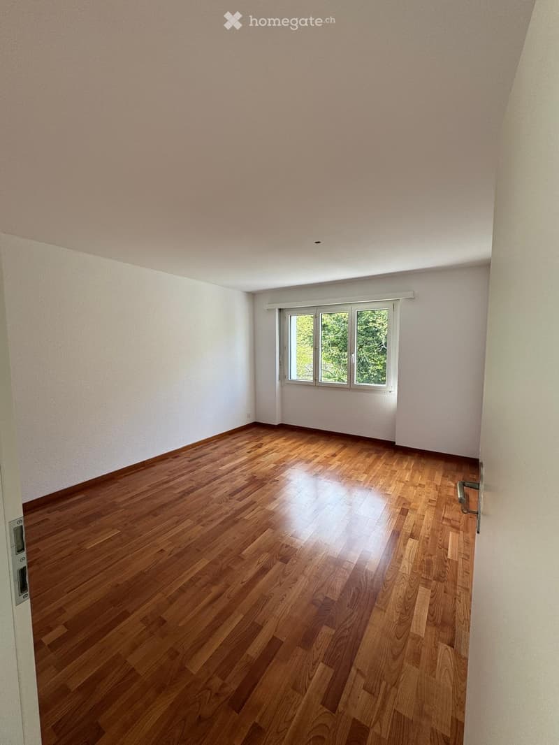 Empty room with wooden flooring and white walls, featuring a large window with blinds and a door visible on the left side.