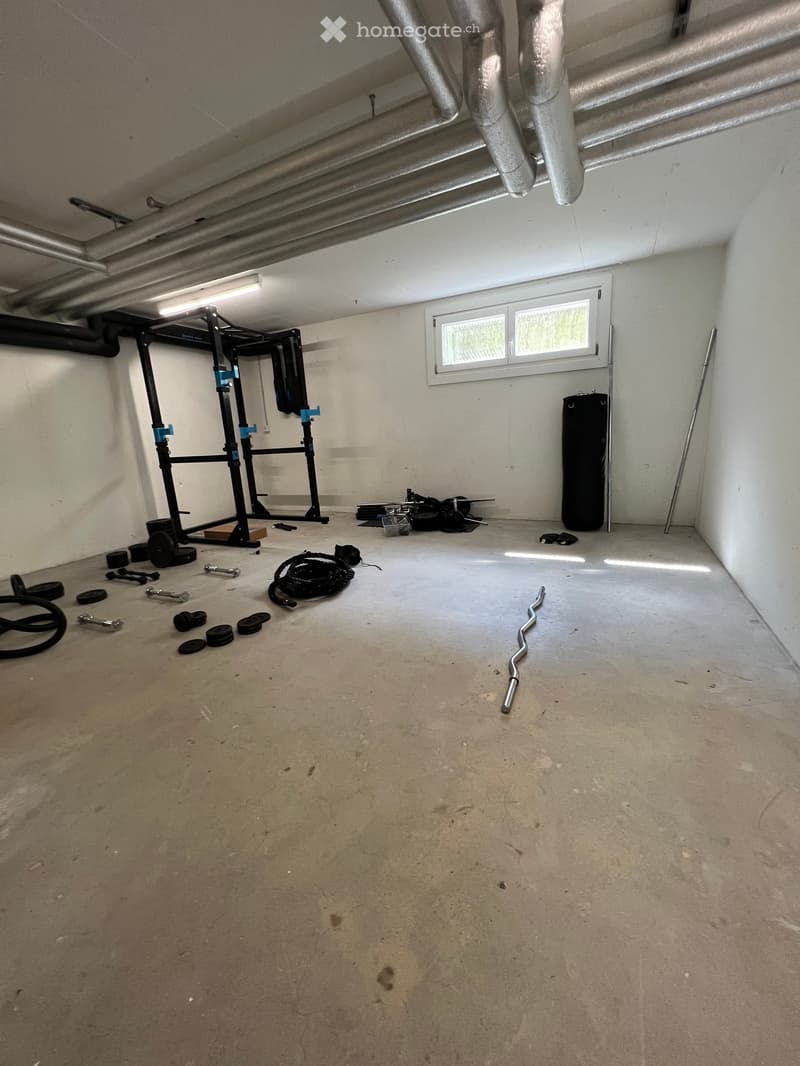 A room with a white wall, a window, a punching bag, a barbell, and a weight bench.