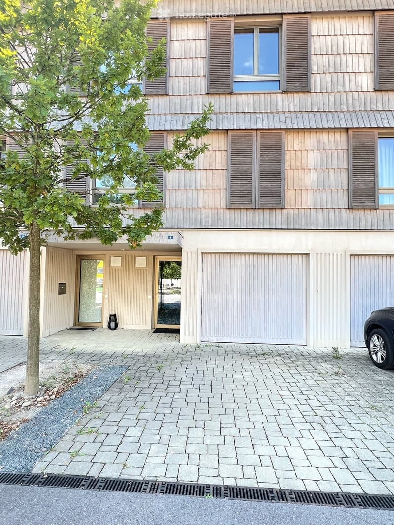 Modern apartment building with wooden exterior, brick walls, large windows, two garages, and a tree in front.
