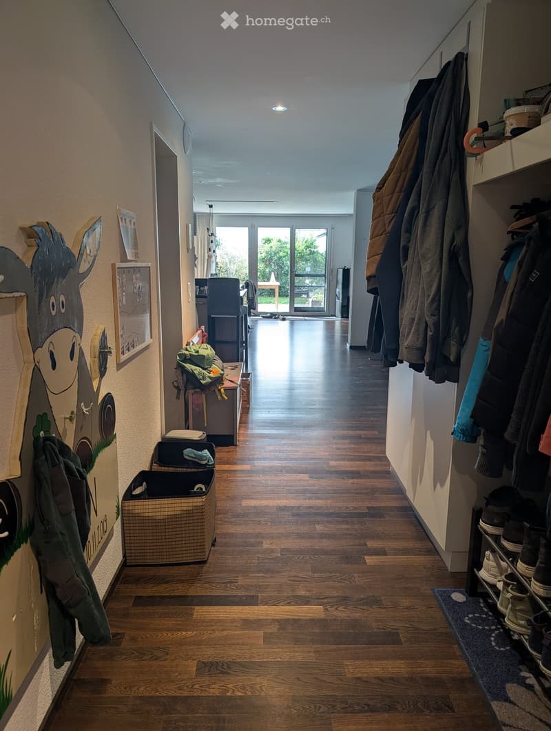 A well-lit hallway with wooden flooring, coat rack, shoe rack, and a small storage basket. The hallway leads to a room with a table and chairs visible in the background.
