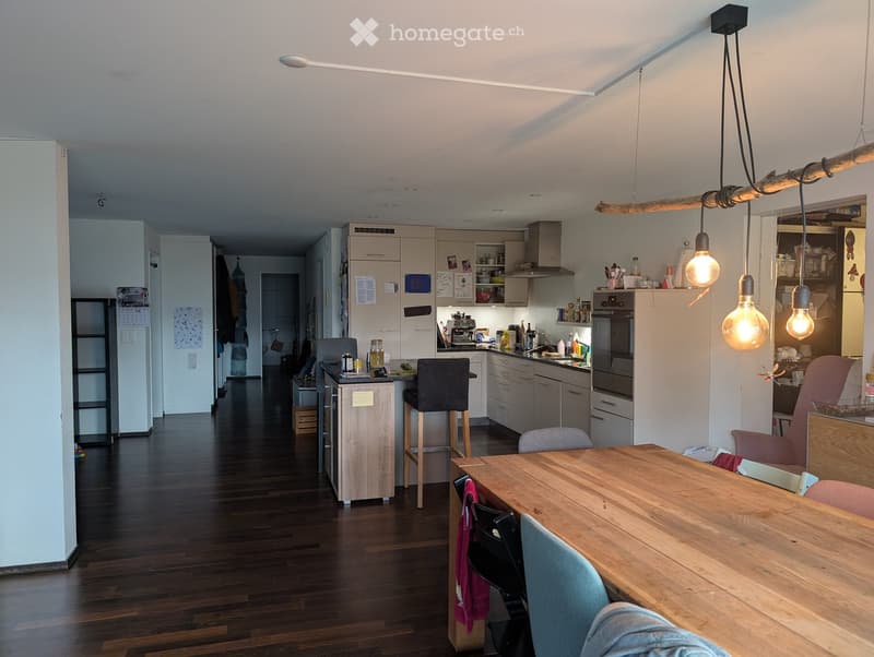 Modern kitchen with wooden table, chairs, stools, and various appliances including a refrigerator and oven. The space features light-colored cabinets and a dark wooden floor.