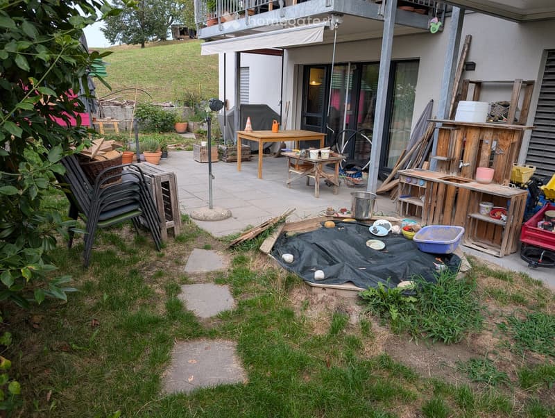 Outdoor patio area with wooden tables, chairs, potted plants, and construction materials, adjacent to a house with a balcony and covered walkway.