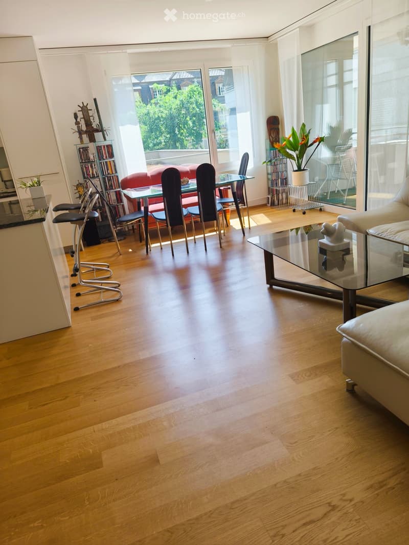 Bright living room with wooden flooring, large windows, a red sofa, a glass coffee table, and a dining area with chairs and a table.
