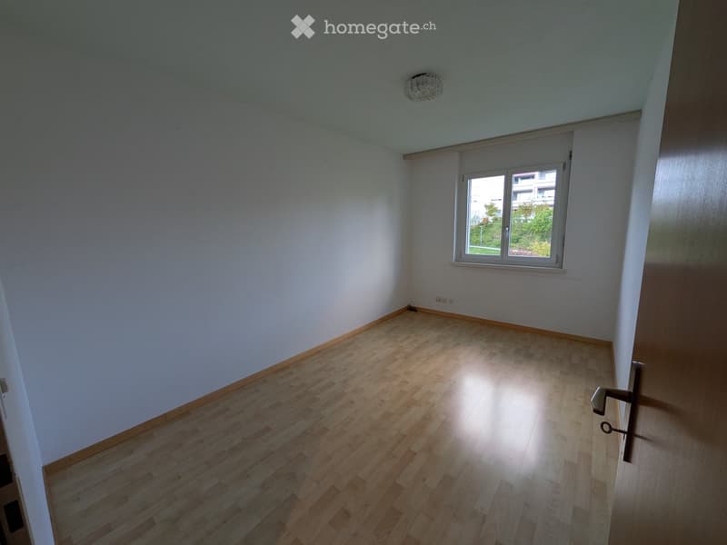Empty room with light wooden flooring, white walls, and a single window with white frame. The ceiling has a light fixture.